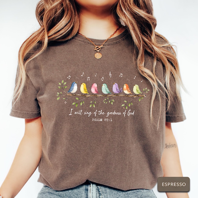 May include: A dark brown short-sleeve t-shirt featuring a graphic of seven colorful birds perched on a leafy branch with musical notes above them. Below the branch, the text reads, I will sing of the goodness of God, PSALM 89:1.