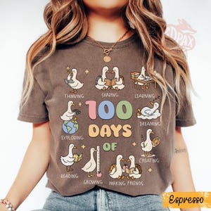 May include: Espresso-colored t-shirt featuring a graphic with cartoon ducks illustrating various activities like reading and creating. The text "100 DAYS OF" is prominently displayed, with additional words like "Thinking" and "Sharing".