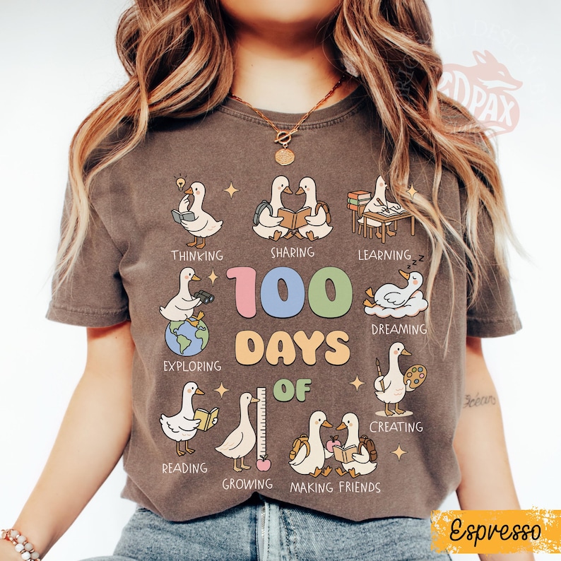 May include: Espresso-colored t-shirt featuring a graphic with cartoon ducks illustrating various activities like reading and creating. The text "100 DAYS OF" is prominently displayed, with additional words like "Thinking" and "Sharing".