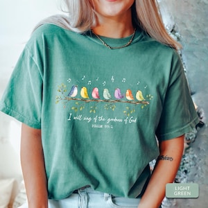 May include: A sage green short-sleeve t-shirt featuring a graphic of seven colorful birds perched on a branch with musical notes above them. Below the birds, the text reads: I will sing of the goodness of God, PSALM 89:1 in white lettering.