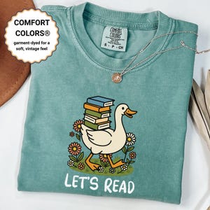 May include: A teal Comfort Colors t-shirt featuring a cartoon goose carrying a stack of books, surrounded by flowers. The text "LET'S READ" is printed below the illustration. The shirt has a soft, vintage feel.