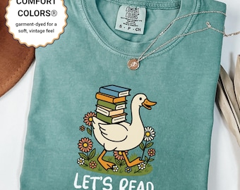 Puede incluir: Una camiseta Comfort Colors de color verde azulado con un dibujo de un ganso de dibujos animados que lleva libros, rodeado de flores. El texto "LET'S READ" está impreso debajo. La camiseta tiene un tacto suave y vintage.