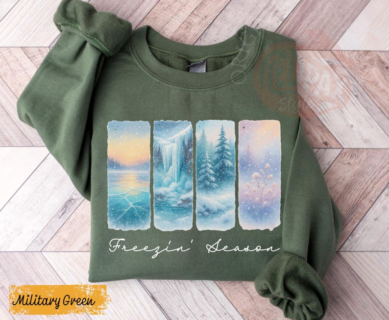 May include: Military green sweatshirt featuring a winter scene design. The design includes four vertical panels depicting a snowy landscape with a frozen lake, waterfall, and evergreen trees. The words "Freezin' Season" are written in white script.