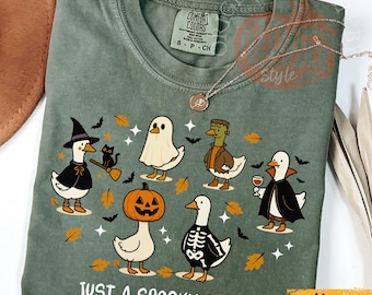 Peut inclure: T-shirt vert mousse Comfort Colors avec un motif sur le thème d'Halloween. Le motif présente des oies de dessin animé déguisées en sorcière, fantôme, Frankenstein, vampire et une oie avec une tête de citrouille. Le texte "JUST A SPOOKY GOOSE" est imprimé en bas.