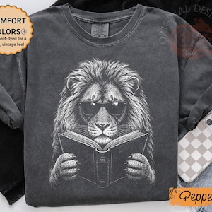 Puede incluir: Una camiseta gris carbón con una ilustración gráfica blanca de un león con gafas de sol leyendo un libro abierto. La camiseta tiene cuello redondo y mangas cortas. Una insignia circular en la parte superior izquierda dice COMFORT COLORS.