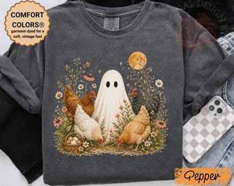 Peut inclure: Un t-shirt gris anthracite avec une impression graphique représentant un fantôme blanc entouré de trois poules, d'un panier d'œufs et de fleurs sauvages sous une pleine lune. Le t-shirt porte une étiquette Comfort Colors et le texte "Original Design by".