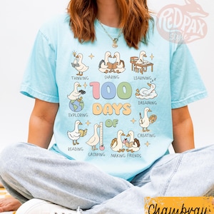 May include: Light blue t-shirt with a graphic featuring cartoon ducks engaged in various activities, including reading and sharing. The text "100 Days of" is prominently displayed. The shirt has a relaxed fit.