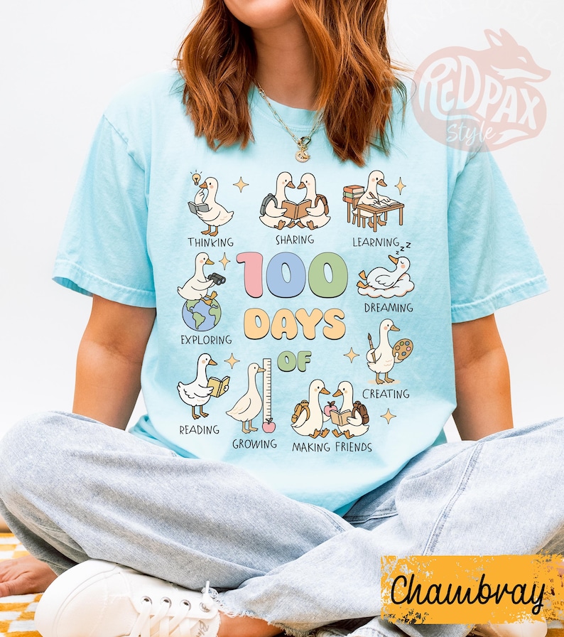 May include: Light blue t-shirt with a graphic featuring cartoon ducks engaged in various activities, including reading and sharing. The text "100 Days of" is prominently displayed. The shirt has a relaxed fit.