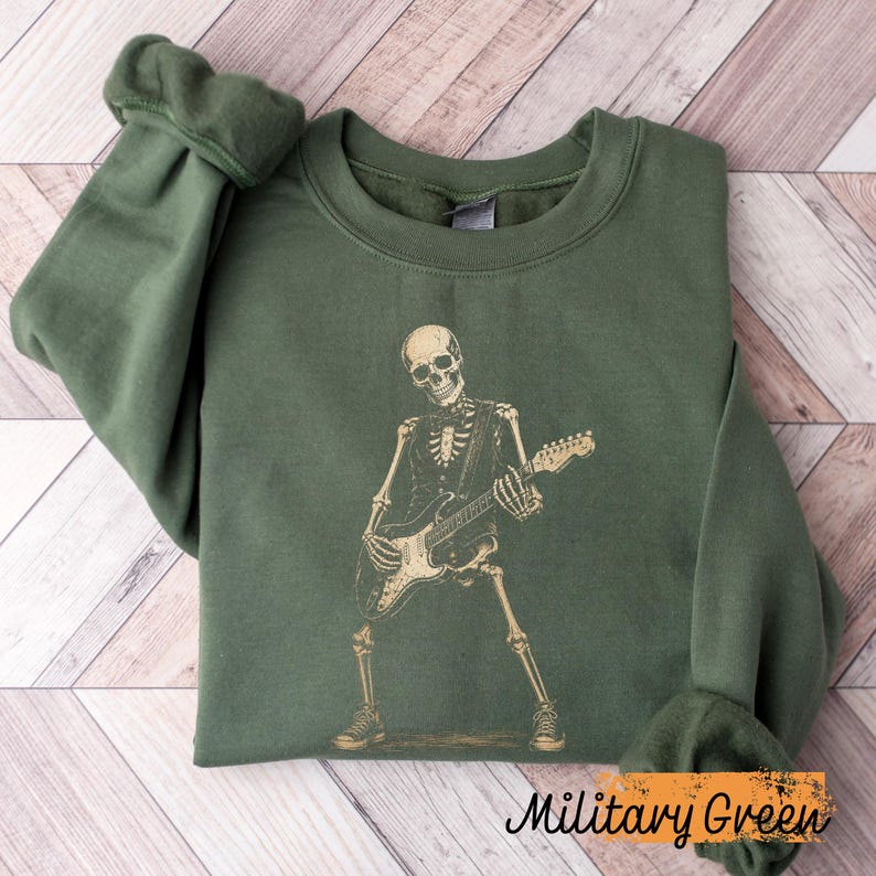 Peut inclure: Un sweat-shirt &agrave; col rond vert olive avec une impression graphique repr&eacute;sentant un squelette jouant de la guitare &eacute;lectrique. Le squelette est repr&eacute;sent&eacute; dans une couleur beige clair, debout dans une pose dynamique tenant l'instrument.