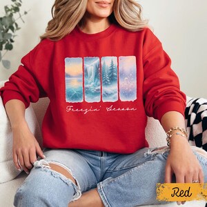 May include: A red sweatshirt featuring a winter landscape design with the text "Freezin' Season". The design includes four rectangular panels depicting snowy scenes with a lake, waterfall, and trees. The sweatshirt is a crew neck style.