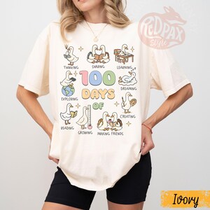May include: Ivory-colored t-shirt featuring a colorful graphic of ducks engaged in various activities, with the text "100 Days Of" and associated words like "Thinking", "Sharing", and "Learning". The shirt has a relaxed fit.