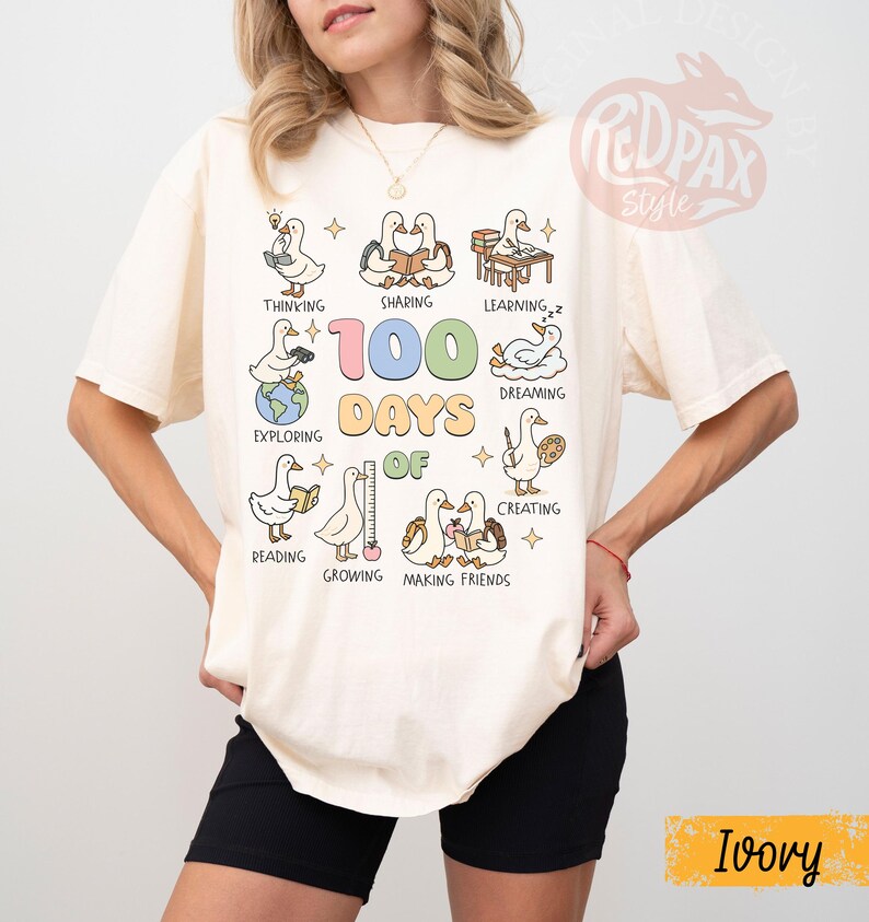 May include: Ivory-colored t-shirt featuring a colorful graphic of ducks engaged in various activities, with the text "100 Days Of" and associated words like "Thinking", "Sharing", and "Learning". The shirt has a relaxed fit.