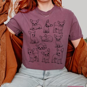 May include: A berry-colored short-sleeve t-shirt featuring a black line-art graphic print of nine different Chihuahua dogs in various poses, including sitting, standing, and lying down. The shirt has a crew neckline and a relaxed fit.