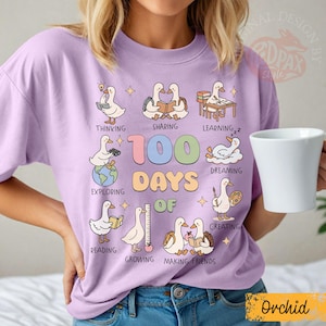 May include: A light purple t-shirt with a graphic design celebrating 100 days of school. The design features cartoon ducks engaged in various activities like reading and exploring. The word "Orchid" is at the bottom.