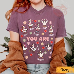 May include: Berry-coloured t-shirt with a design of cartoon geese and positive affirmations. The geese are surrounded by hearts and flowers, with phrases like "Brave," "Kind," and "Unique." The text "YOU ARE" is prominently displayed.