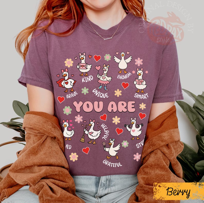 May include: Berry-coloured t-shirt with a design of cartoon geese and positive affirmations. The geese are surrounded by hearts and flowers, with phrases like "Brave," "Kind," and "Unique." The text "YOU ARE" is prominently displayed.