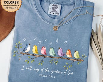 May include: A blue jean colored t-shirt featuring a graphic of seven colorful birds perched on a branch with musical notes. The text reads, I will sing of the goodness of God, PSALM 89:1. The shirt has a Comfort Colors brand label at the neck.
