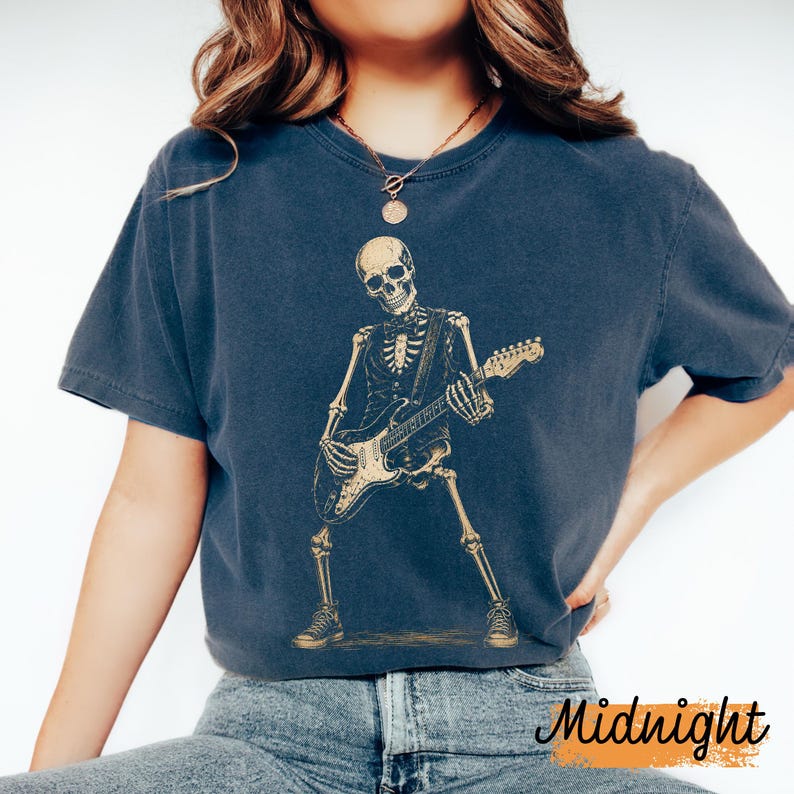 Peut inclure: Un t-shirt bleu fonc&eacute; &agrave; manches courtes avec une impression graphique repr&eacute;sentant un squelette portant un gilet et un n&oelig;ud papillon tout en jouant de la guitare &eacute;lectrique. Le squelette est en beige clair, dans une pose dynamique.