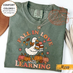 May include: Moss green Comfort Colors t-shirt with a graphic that reads "Fall in Love with Learning." The design features a white duck holding books and an apple, surrounded by pumpkins and fall leaves.