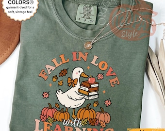 May include: Moss green Comfort Colors t-shirt with a graphic that reads "Fall in Love with Learning." The design features a white duck holding books and an apple, surrounded by pumpkins and fall leaves.