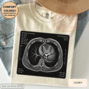 Op de afbeelding: Ivoor kleurig T-shirt met een afbeelding van een kat in een borstscan, met een hart. Het shirt heeft de tekst "COMFORT COLORS® garment-dyed for a soft, vintage feel" en "Soft Ink Print (DTG)".
