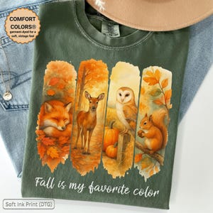 Könnte beinhalten: Ein olivgrünes T-Shirt mit einem Grafikdruck aus vier vertikalen Feldern, die einen schlafenden Fuchs, ein Reh, eine Eule und ein Eichhörnchen zwischen Herbstblättern und Kürbissen zeigen. Der Text "Fall is my favorite color" ist in Weiß unter dem Motiv gedruckt.