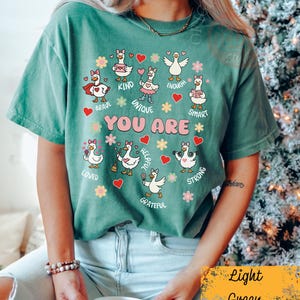 May include: Light green t-shirt featuring cartoon geese with positive affirmations. The text "YOU ARE" is central, surrounded by words such as "BRAVE", "KIND", and "UNIQUE". The shirt has a relaxed fit and is a comfortable style.