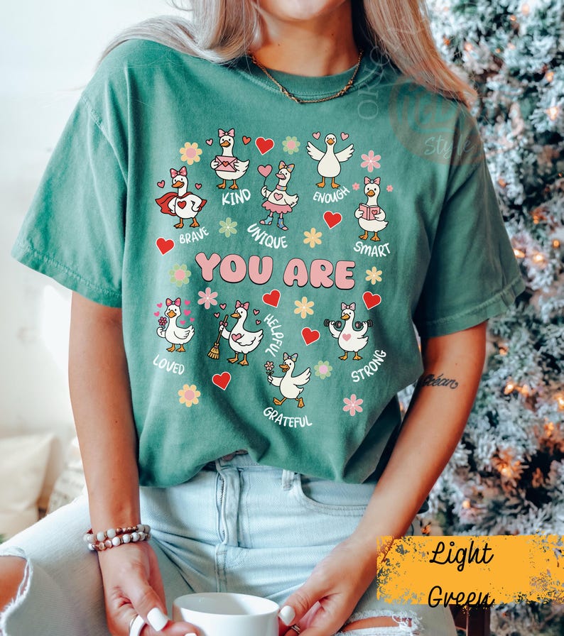 May include: Light green t-shirt featuring cartoon geese with positive affirmations. The text "YOU ARE" is central, surrounded by words such as "BRAVE", "KIND", and "UNIQUE". The shirt has a relaxed fit and is a comfortable style.