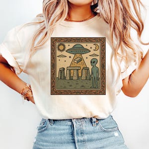 Op de afbeelding: Beige T-shirt met een ingelijste afbeelding van een UFO boven Stonehenge, een alien, zon, maan en sterren. Het ontwerp heeft een bruine rand. Het shirt is gemaakt van een zacht materiaal.