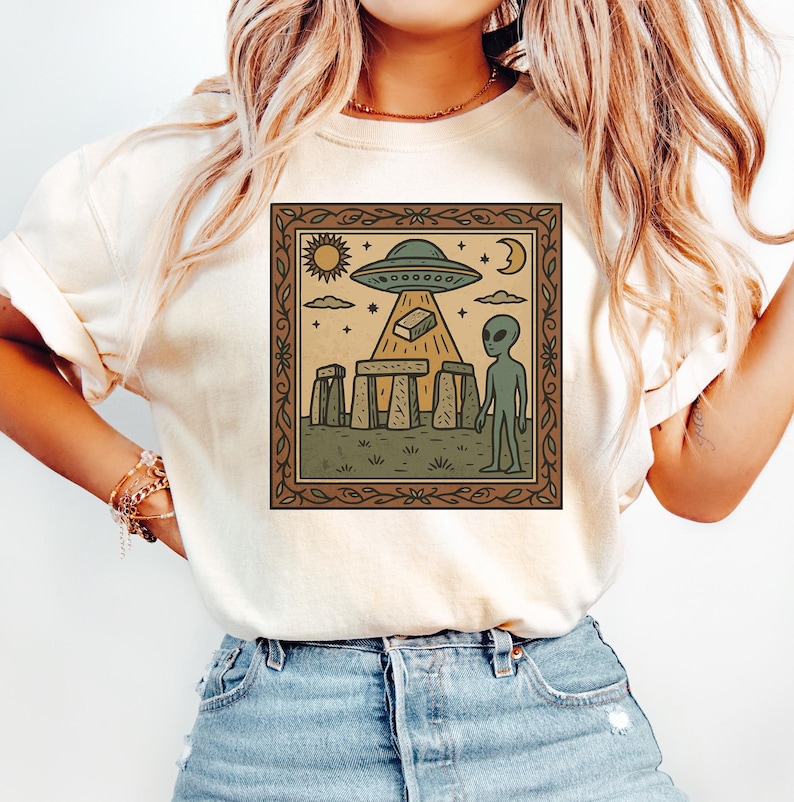 Op de afbeelding: Beige T-shirt met een ingelijste afbeelding van een UFO boven Stonehenge, een alien, zon, maan en sterren. Het ontwerp heeft een bruine rand. Het shirt is gemaakt van een zacht materiaal.