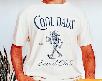 Könnte beinhalten: Elfenbeinfarbenes T-Shirt mit marineblauem Aufdruck. Der Aufdruck zeigt den Text "Cool Dads Social Club" und ein Skelett mit Mütze, das raucht und einen Bierkrug hält. Das Jahr "2024" ist ebenfalls enthalten.