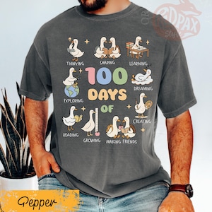 May include: A dark gray t-shirt with a colorful graphic celebrating "100 Days of" school. The design features cartoon geese engaged in various activities like reading, sharing, and creating. The word "Pepper" is at the bottom.
