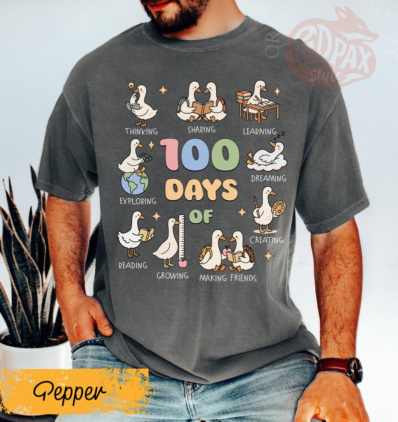 May include: A dark gray t-shirt with a colorful graphic celebrating "100 Days of" school. The design features cartoon geese engaged in various activities like reading, sharing, and creating. The word "Pepper" is at the bottom.