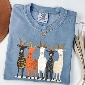 May include: A blue Comfort Colors t-shirt featuring a graphic of five cartoon goats in various colors. The goats have large horns and are standing side-by-side. A gold necklace with a pendant is draped across the shirt.