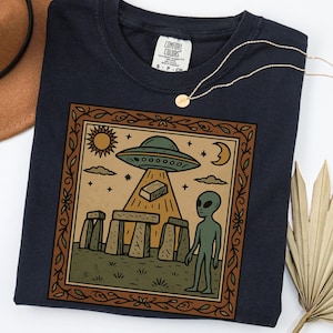 Op de afbeelding: Marineblauwe Comfort Colors T-shirt met een ingelijste afbeelding van een UFO, alien, Stonehenge, zon en maan. Een gouden ketting met een kleine hanger ligt over het T-shirt.