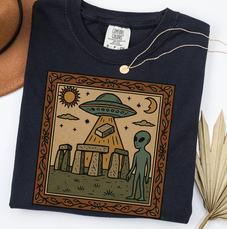 Op de afbeelding: Marineblauwe Comfort Colors T-shirt met een ingelijste afbeelding van een UFO, alien, Stonehenge, zon en maan. Een gouden ketting met een kleine hanger ligt over het T-shirt.