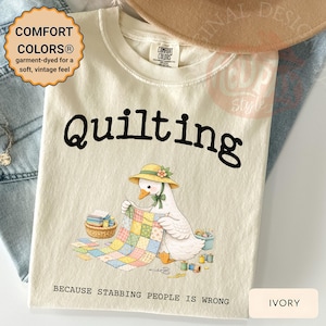 May include: Ivory-colored t-shirt with the word "Quilting" in black. A cartoon goose wearing a hat is depicted quilting. The text "BECAUSE STABBING PEOPLE IS WRONG" is below the image. The shirt has a vintage feel.