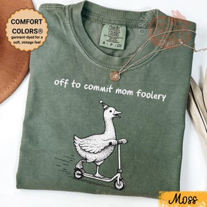 May include: A moss green t-shirt with the slogan "off to commit mom foolery". The graphic shows a goose on a scooter. The shirt is garment-dyed for a soft, vintage feel. The t-shirt has a Comfort Colors label.