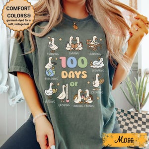 May include: Moss green t-shirt with a graphic celebrating 100 days of school. The design features cartoon ducks engaged in various activities, with the text "100 Days of" and other words. The shirt also has the text "COMFORT COLORS&reg;" and "Moss".