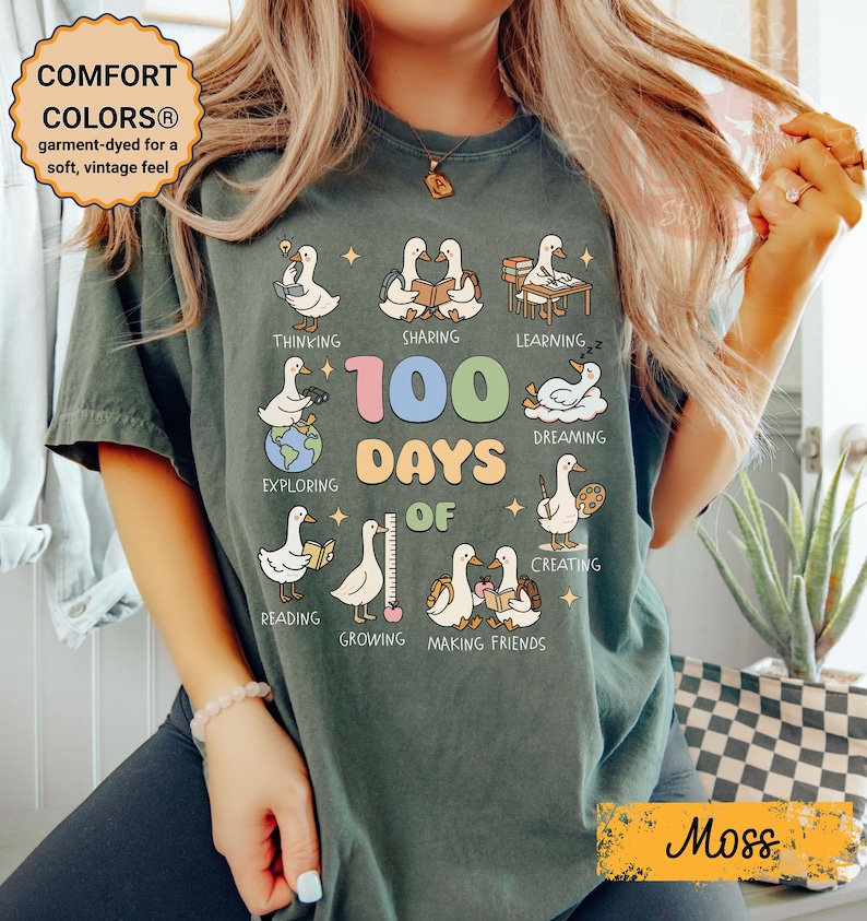 May include: Moss green t-shirt with a graphic celebrating 100 days of school. The design features cartoon ducks engaged in various activities, with the text "100 Days of" and other words. The shirt also has the text "COMFORT COLORS&reg;" and "Moss".