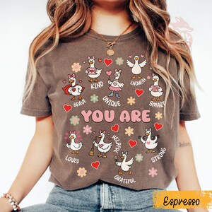 May include: Espresso-coloured t-shirt with a graphic of cartoon geese and positive affirmations such as "Brave," "Kind," and "Unique." The design includes hearts and flowers, with the words "YOU ARE" displayed. This is a casual wear item.