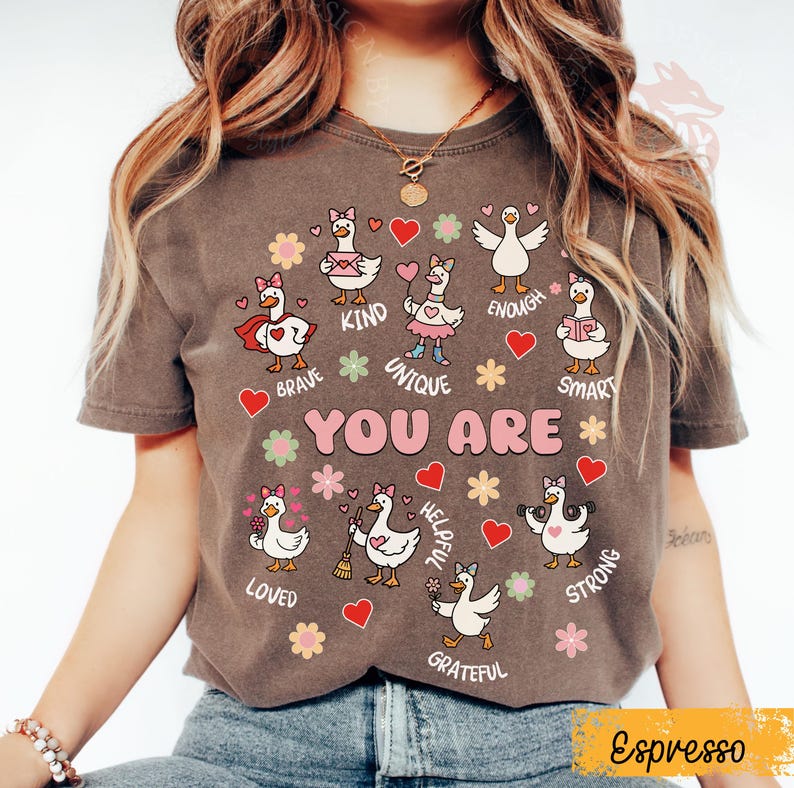 May include: Espresso-coloured t-shirt with a graphic of cartoon geese and positive affirmations such as "Brave," "Kind," and "Unique." The design includes hearts and flowers, with the words "YOU ARE" displayed. This is a casual wear item.