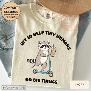 Puede incluir: Camiseta color marfil con un diseño de mapache montado en un patinete azul y sosteniendo una bebida. El texto dice "OFF TO HELP TINY HUMANS DO BIG THINGS". La camiseta está teñida para una sensación suave y vintage.