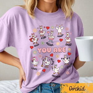 May include: A lilac t-shirt with cartoon geese and positive affirmations. The geese are decorated with hearts and flowers, with phrases such as "Brave," "Kind," and "Unique." The words "YOU ARE" are in the center. The word "Orchid" is at the bottom.