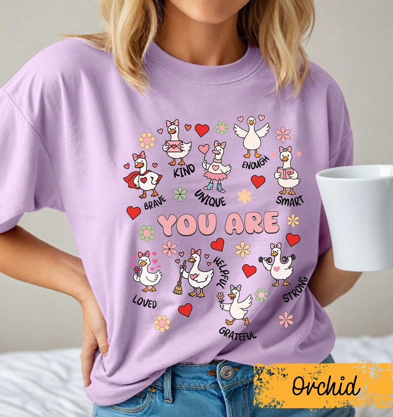May include: A lilac t-shirt with cartoon geese and positive affirmations. The geese are decorated with hearts and flowers, with phrases such as "Brave," "Kind," and "Unique." The words "YOU ARE" are in the center. The word "Orchid" is at the bottom.