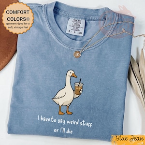 May include: Blue jean Comfort Colors t-shirt featuring a goose graphic holding an iced coffee. The text "I have to say weird stuff or I'll die" is printed below the goose. A gold necklace is draped across the shirt.