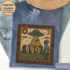 Op de afbeelding: Blauw spijkerbroekkleurig T-shirt met een ingelijste afbeelding met een UFO, alien, Stonehenge, zon en maan. Het shirt heeft de tekst "COMFORT COLORS&reg; garment-dyed for a soft, vintage feel" en "BLUE JEAN".