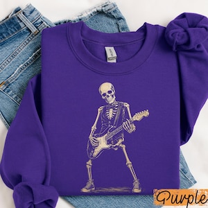 Peut inclure: Un sweat-shirt &agrave; col rond violet avec un motif cr&egrave;me repr&eacute;sentant un squelette portant un gilet et un n&oelig;ud papillon tout en jouant de la guitare &eacute;lectrique. Le motif est centr&eacute; sur le devant du v&ecirc;tement, pos&eacute; sur un tissu en jean bleu.