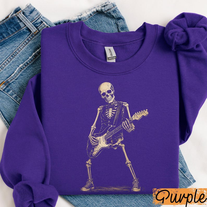 Peut inclure: Un sweat-shirt &agrave; col rond violet avec un motif cr&egrave;me repr&eacute;sentant un squelette portant un gilet et un n&oelig;ud papillon tout en jouant de la guitare &eacute;lectrique. Le motif est centr&eacute; sur le devant du v&ecirc;tement, pos&eacute; sur un tissu en jean bleu.