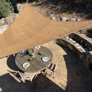 May include: A triangular tan woven shade sail suspended over a circular wooden outdoor dining table. The table is set with plates, a vase of flowers, and a glass. The shade sail features a textured, rope-like weave and reinforced edges.